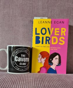 Boek Lover birds van Leanne Egan tegen een roze achtergrond. Links van het boek staat een mok van de Cavern Club.