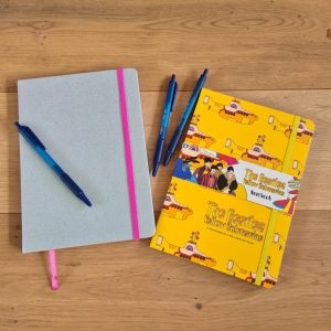 Verhalen in mijn hoofd: 2 schriften op een houten vloer met daarbij 3 blauwe pennen. Het linker schrift is zilverigglitter met een roze elastiek en leeslint, het rechter schrift is bedrukt met de yellow submarine  van The Beatles. Erop staat The Beatles Yellow Submarine. Het heeft een geel elastiek eromheen en een papieren wikkel waarop de getekende Beatles staan.
