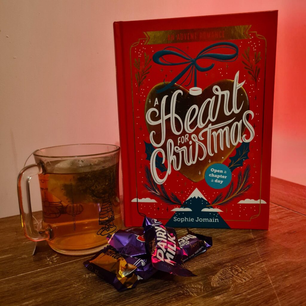 Rechts staat het boek a Heart for Christmas (rode kaft) met daar links van een kop thee. Voor het boek liggen chocolaatjes. De achtergrond is een beetje oranjig, de ondergrond is van hout | 3 must-have adventskalenders voor boekenwurmen