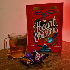 Rechts staat het boek a Heart for Christmas (rode kaft) met daar links van een kop thee. Voor het boek liggen chocolaatjes. De achtergrond is een beetje oranjig, de ondergrond is van hout | 3 must-have adventskalenders voor boekenwurmen