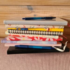 Stapel gekleurde notitieboekjes met daarop 2 blauwe pennen. De ondergrond is een roze corduroy stof en de achtergrond een wand van houten planken.