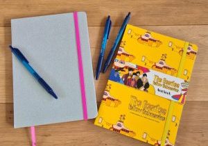 Verhalen in mijn hoofd: 2 schriften op een houten vloer met daarbij 3 blauwe pennen. Het linker schrift is zilverigglitter met een roze elastiek en leeslint, het rechter schrift is bedrukt met de yellow submarine van The Beatles. Erop staat The Beatles Yellow Submarine. Het heeft een geel elastiek eromheen en een papieren wikkel waarop de getekende Beatles staan.